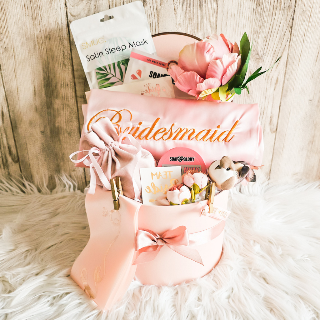 Bridesmaid gift basket with pink satin bag, flowers, and other items on a fluffy surface.